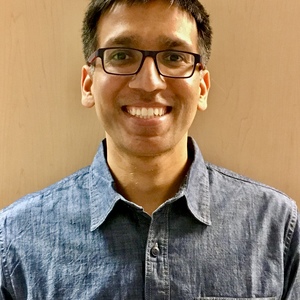 image d'un chercheur portant des lunettes et u bouton bleu, debout devant un mur uni