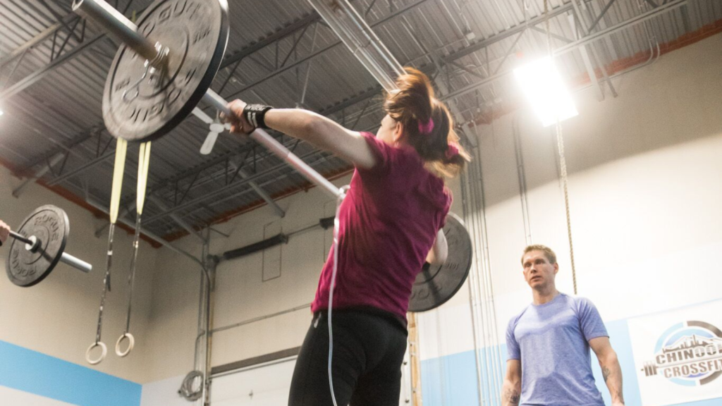 Nicki Perkins dans une salle de sport Crossfit, soulevant une haltère avec un coach qui l'encourage à faire l'exercice.